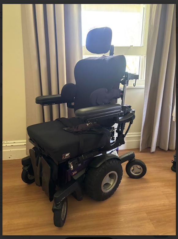 A motorised wheelchair with a padded seat, backrest, headrest and adjustable armrests sits indoors on a wooden floor near a window with beige curtains. The chair has a right‑side joystick controller, large rear wheels, small front casters, and a “JAY” branded seat cushion