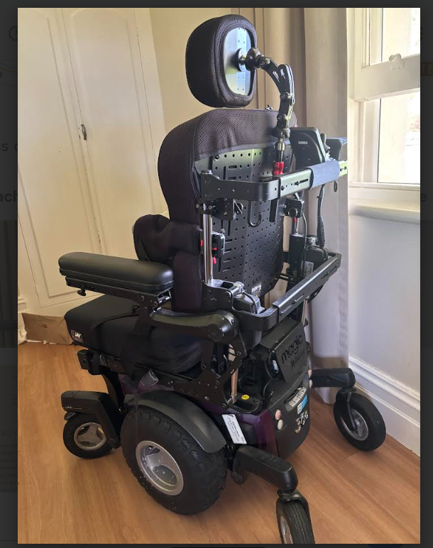 A powered wheelchair with a padded JAY seating system, adjustable armrests and a right‑side joystick controller sits on a wooden floor near a window with beige curtains. The mobility chair features large rear wheels and small front casters, designed for indoor accessibility and user comfort