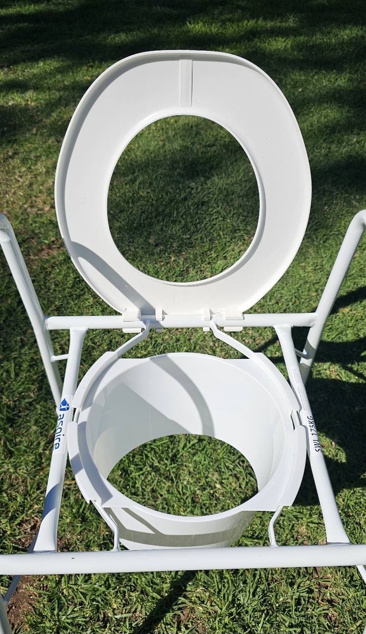 White portable commode chair with armrests and open seat, shown outdoors on grass to display frame and condition